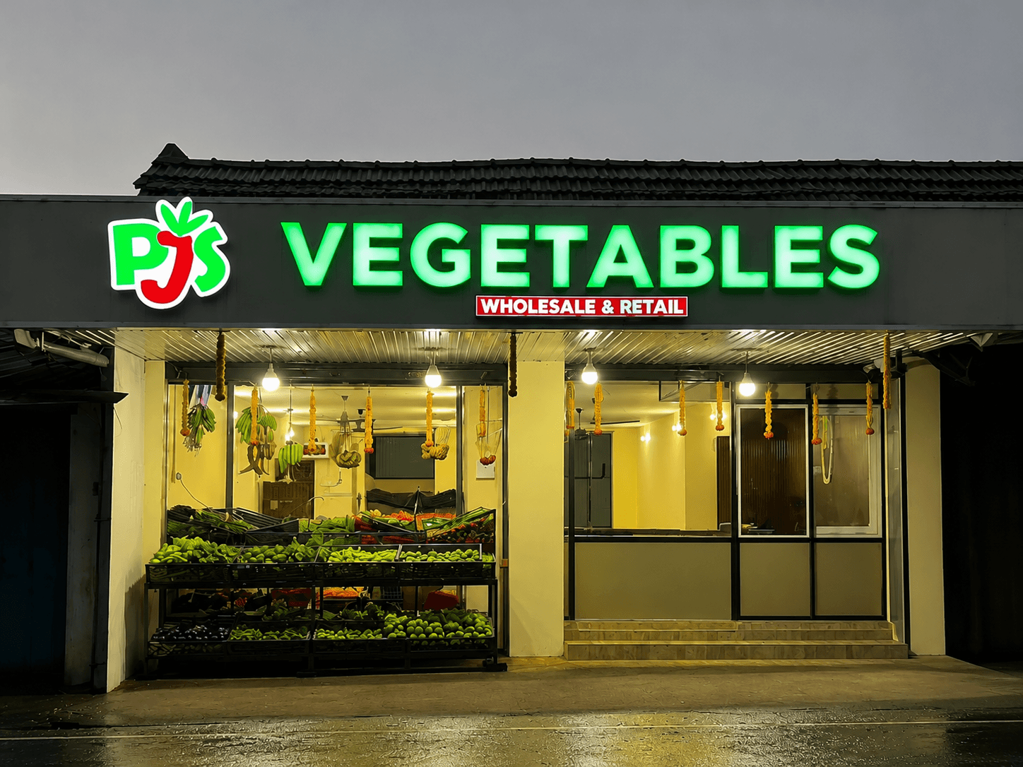 The PJS Vegetables storefront in Changanacherry, lit up in the evening with fresh produce on display and a marigold garland over the entrance
